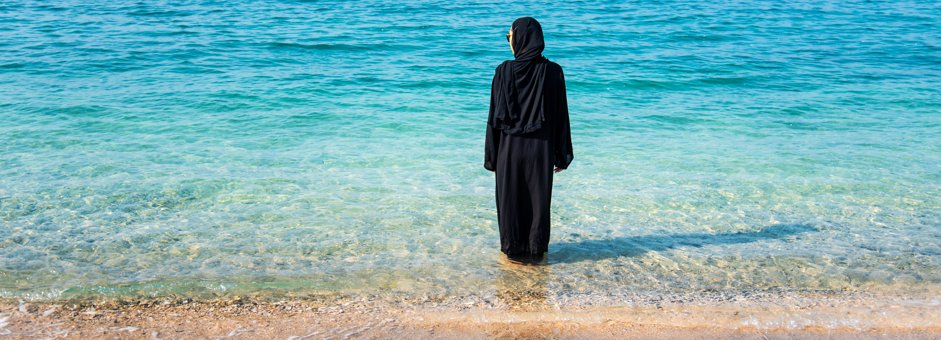 Modest woman in a black abaya on the beach, feet in the water, gazing at the beautiful blue sea with her back to the camera.